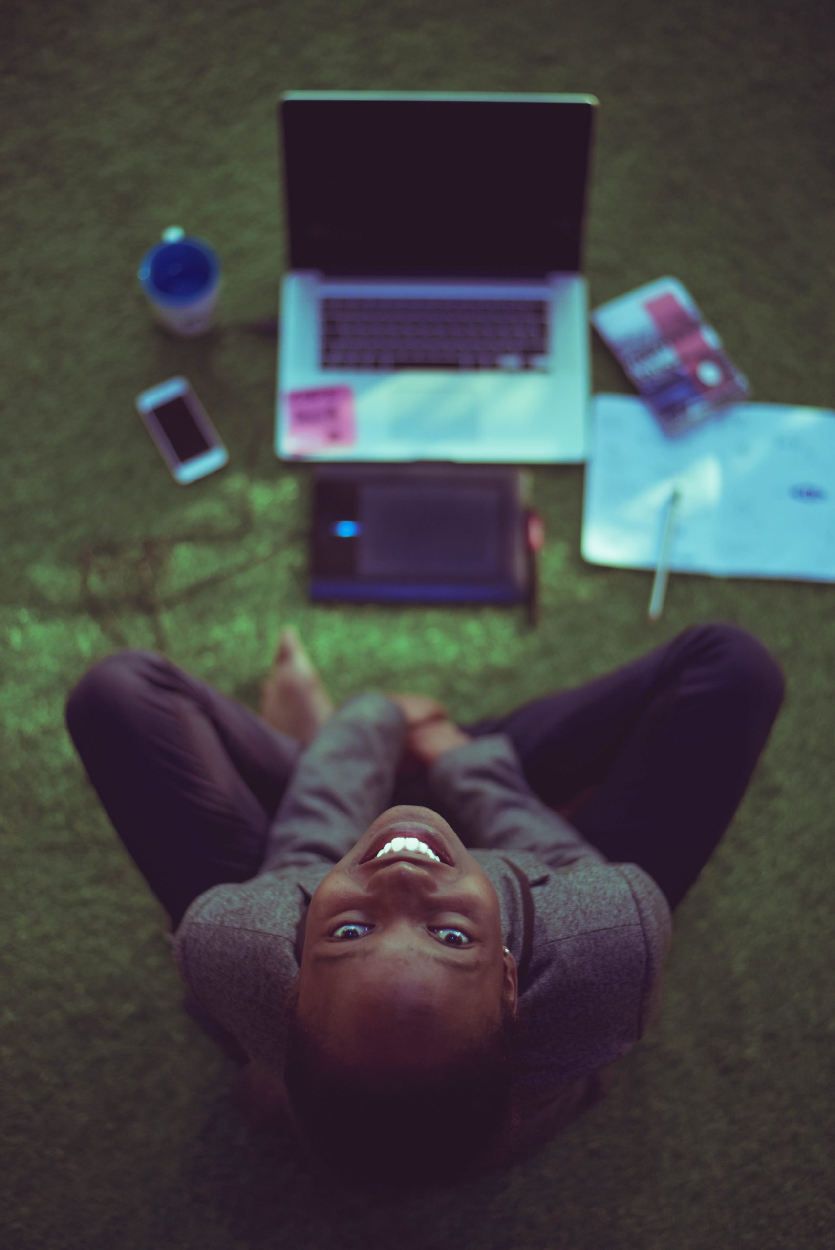 A beautiful, young woman looking up from her workspace, smiling. Workspace is a table with a laptop, phone, mug, & calendar spread out on grass as she is outside.